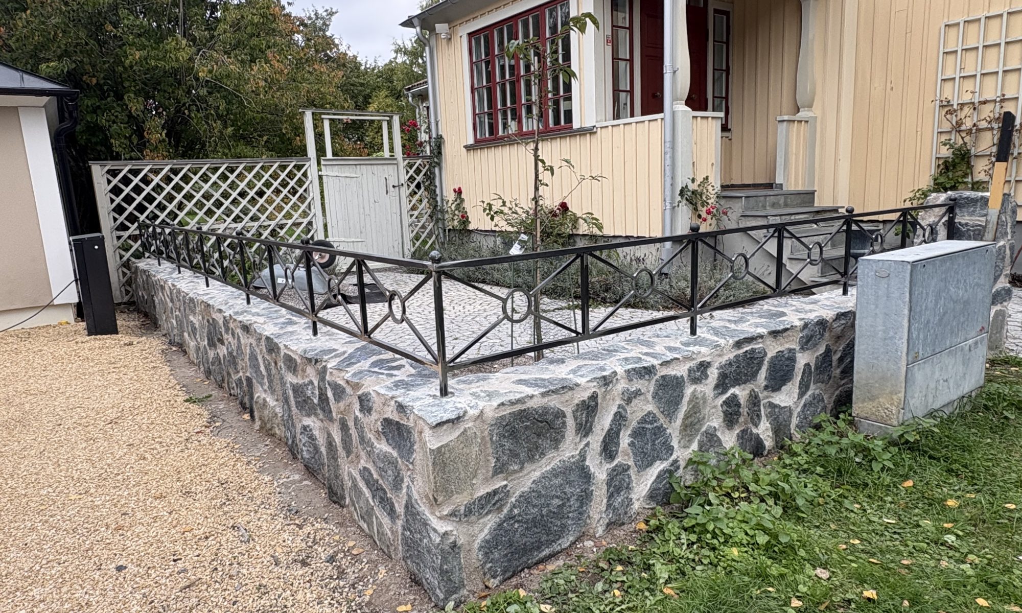 Svart lågräcke med kryss, ringar och kulor på en stenmur framför en gul villa i Bromma.